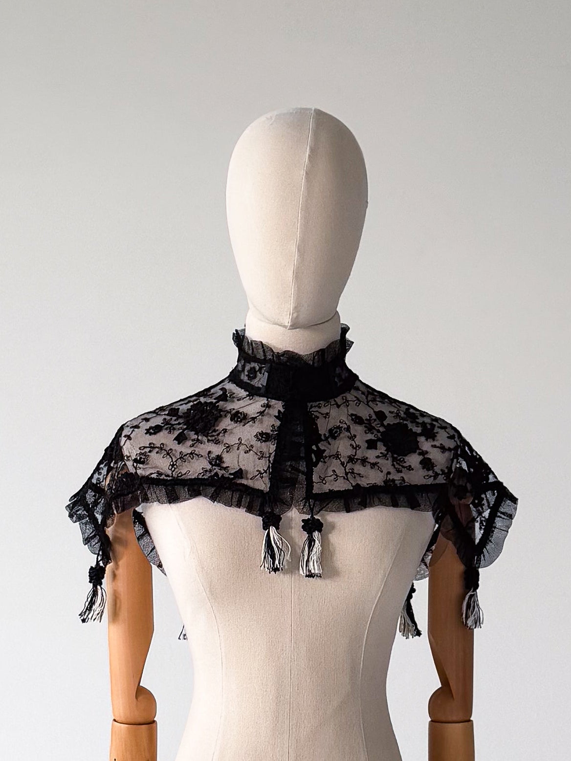 front view of the black lace shawl styled on a dress form, with a ruffled collar closing at the neck and tassel fringe