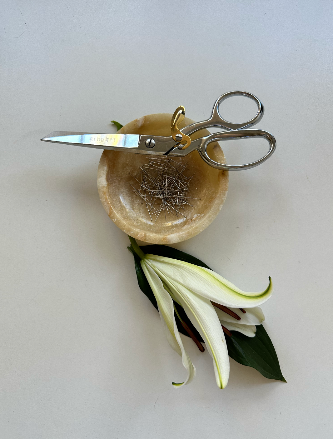 A pair of silver Gingher sewing shears rests on a small marble bowl filled with pins, with a white lily and green leaves placed below on a light surface—an elegant still life evoking craftsmanship and calm precision.