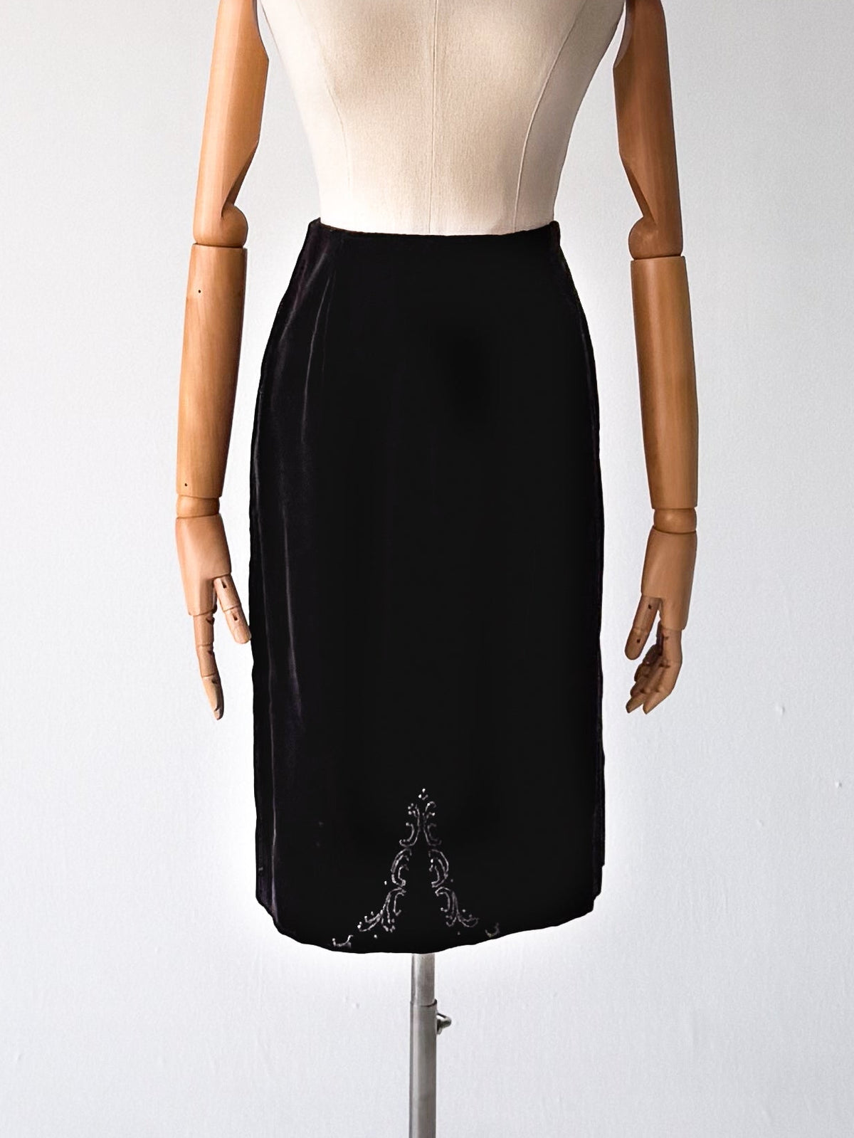 A black knee-length skirt displayed on a mannequin dress form with wooden arms, standing on a metal tripod stand against a plain light gray studio background.
