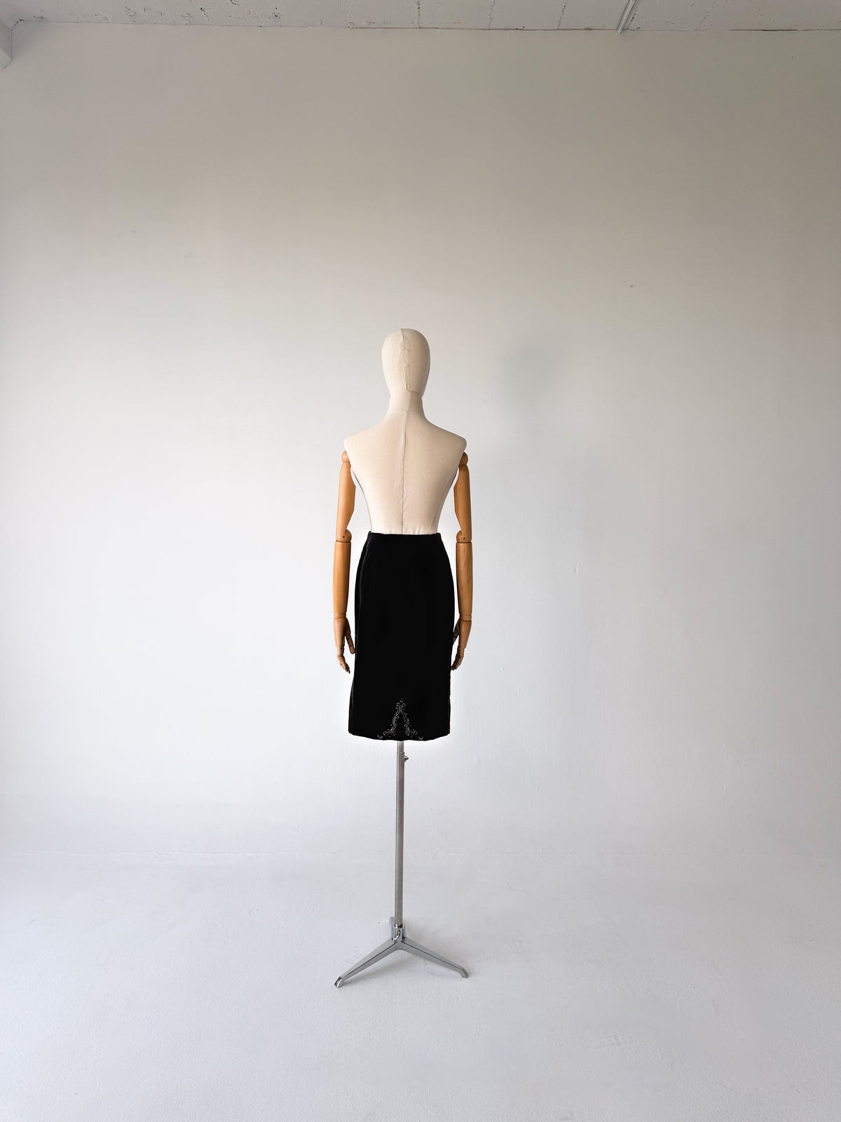 A black knee-length skirt displayed on a dress form mannequin with wooden arms, standing on a silver tripod stand against a plain white studio background.