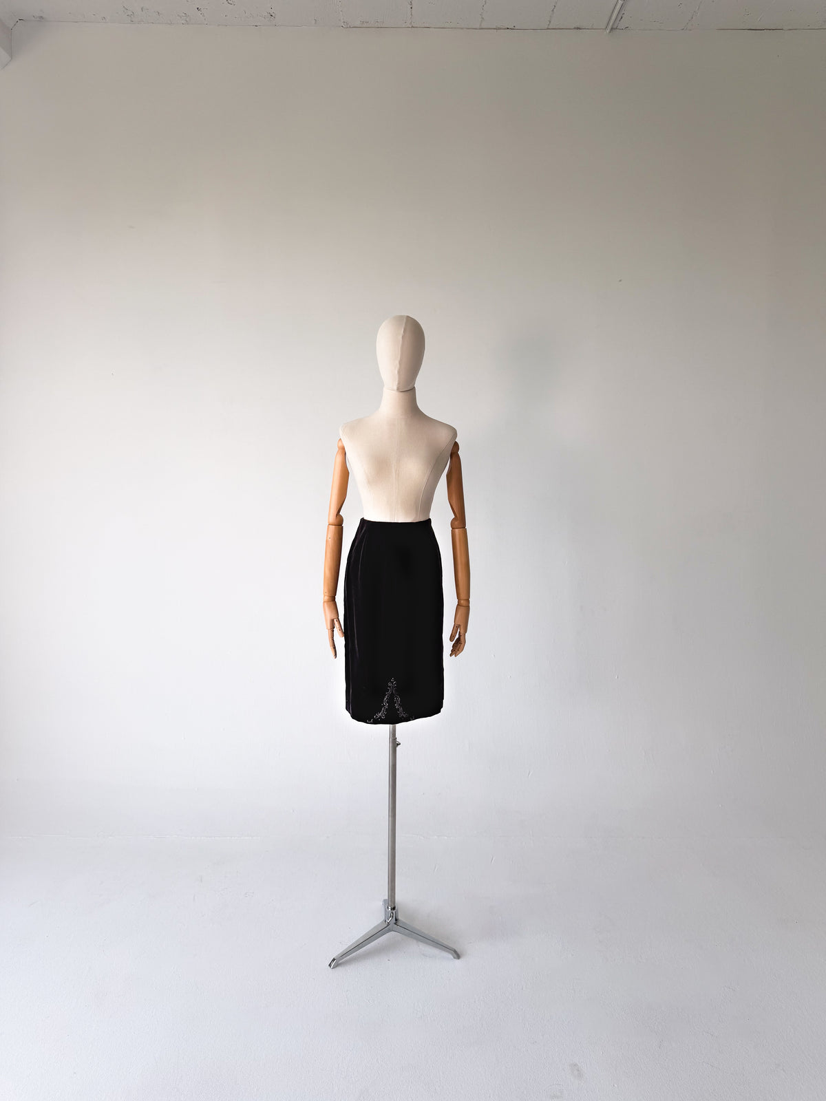A black knee-length skirt displayed on a mannequin dress form with wooden arms, standing on a metal tripod stand against a plain light gray studio background.