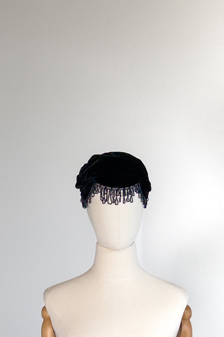 A black velvet hat with a ruffled edge and dangling tassel-like trim, displayed on a cream mannequin head against a plain light gray background.