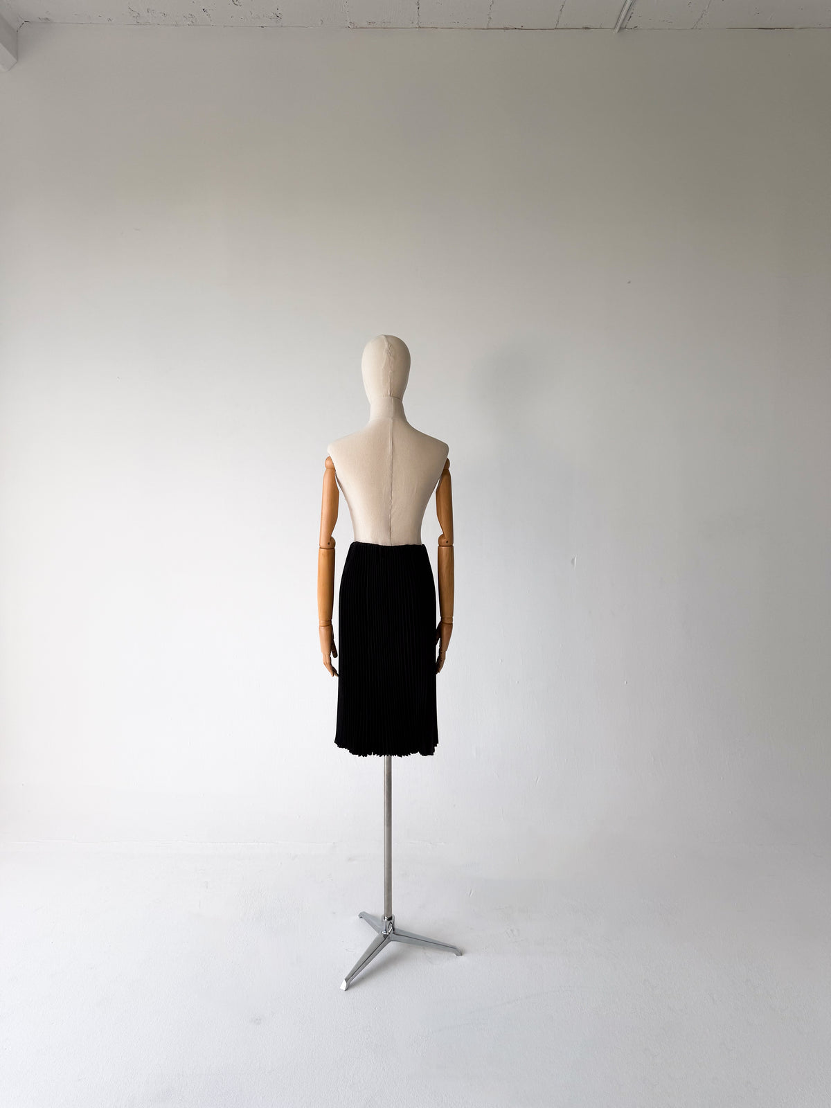 A black midi skirt displayed on a wooden mannequin from the back, standing on a silver metal tripod stand against a plain white studio background.