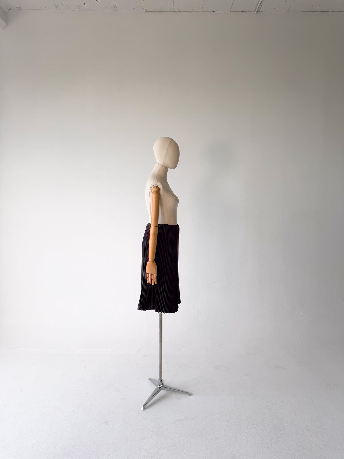 A black pleated skirt displayed on a headless mannequin torso with a wooden arm, standing on a metal tripod stand against a plain light gray studio background.