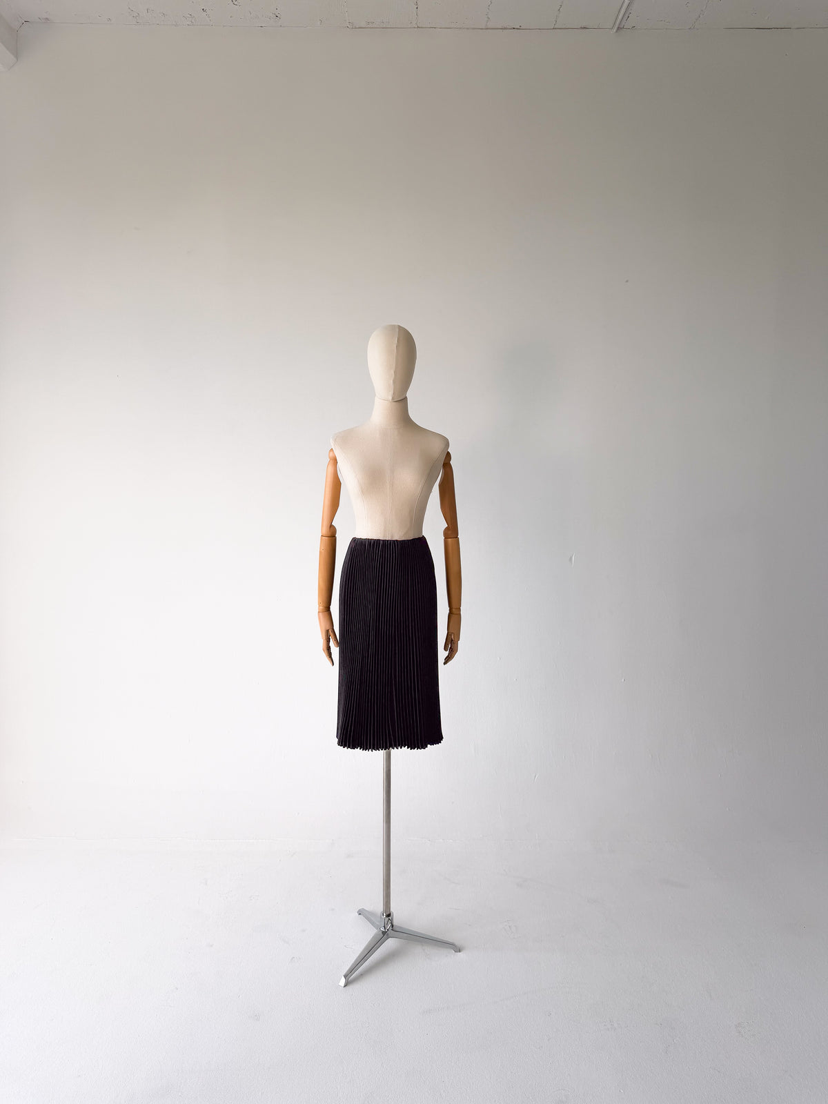 A black pleated midi skirt displayed on a torso mannequin with wooden arms, standing on a metal tripod stand against a plain white studio background.