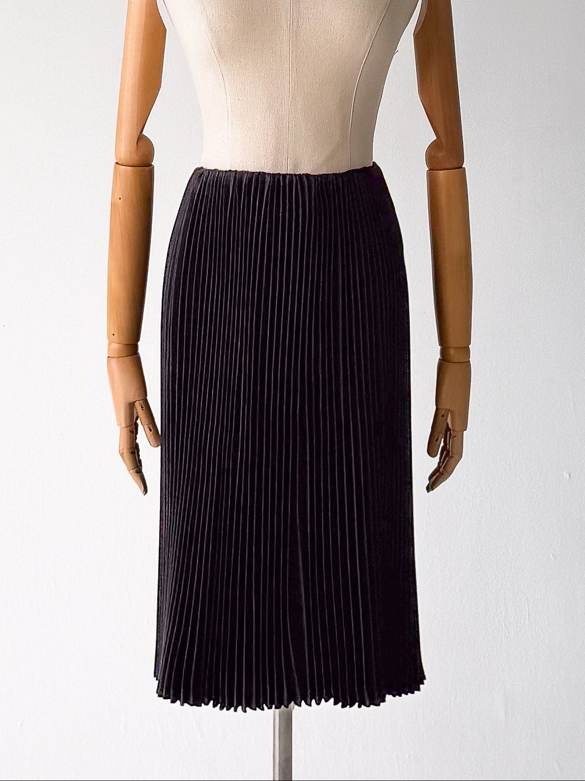 A pleated midi skirt in very dark navy or near-black fabric, displayed on a beige mannequin with wooden arms against a plain light gray background.