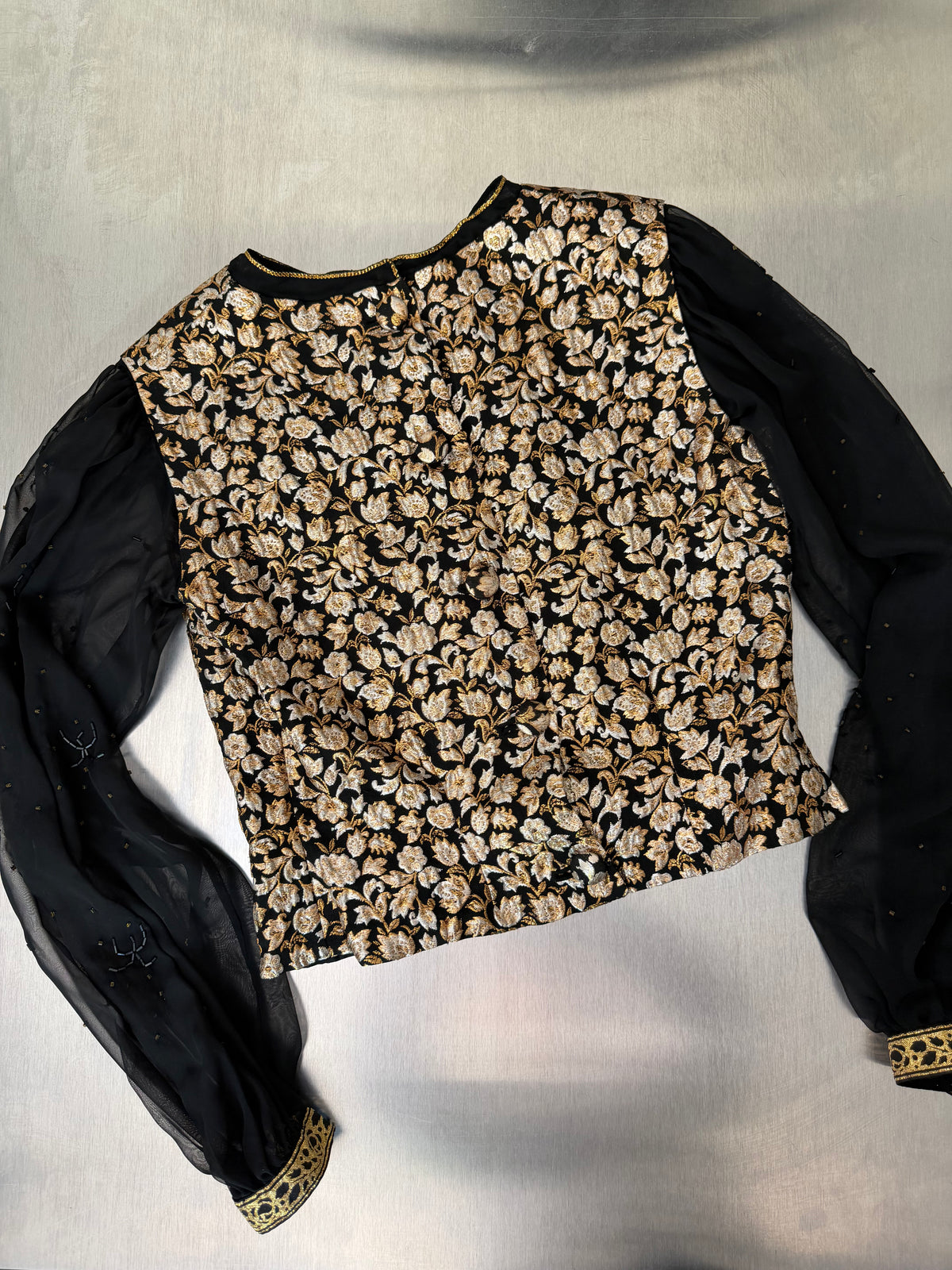 gold brocade blouse laid flat, front view, showing the sheer sleeves with gold tape trim at the cuffs