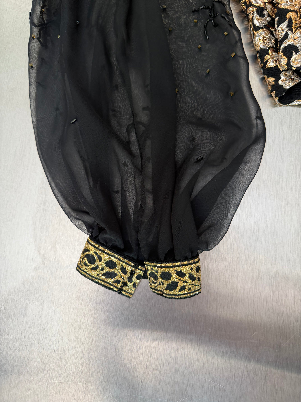 both sheer black sleeves of the gold brocade blouse, gathered at the cuff with gold scrollwork tape trim
