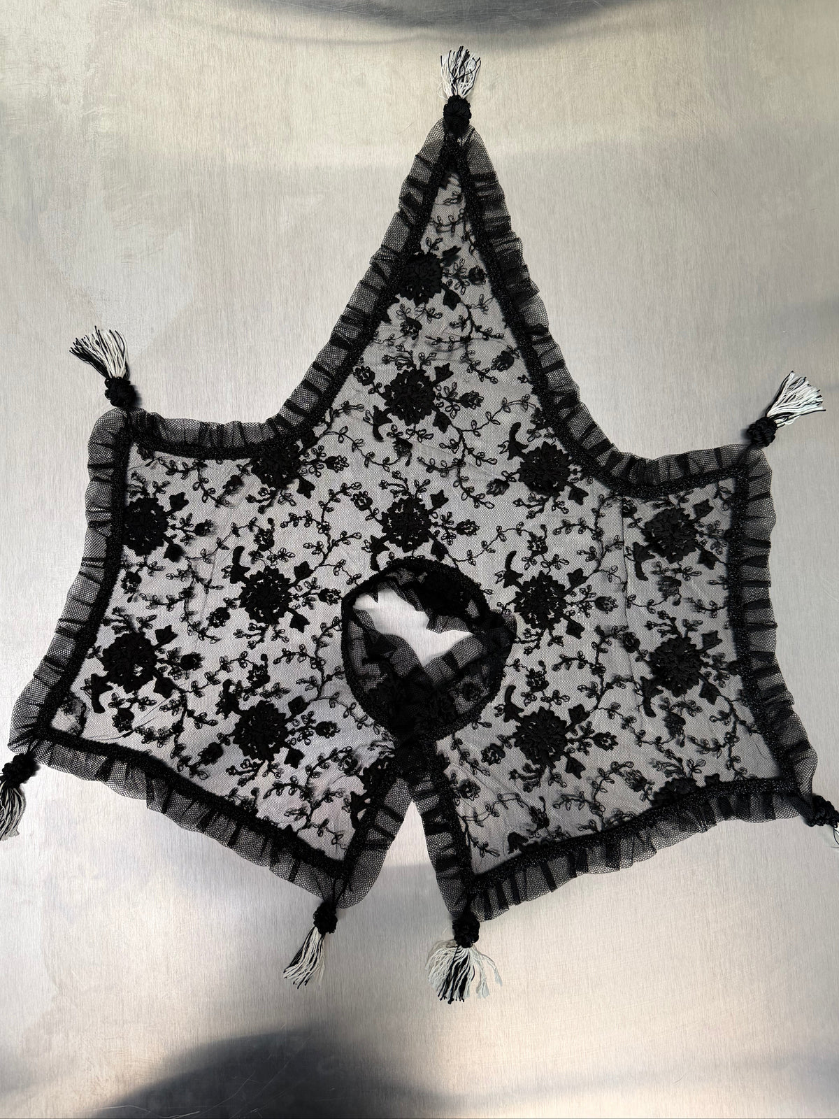 black lace shawl spread flat from above, showing the neck opening and star-shaped silhouette with tasseled points