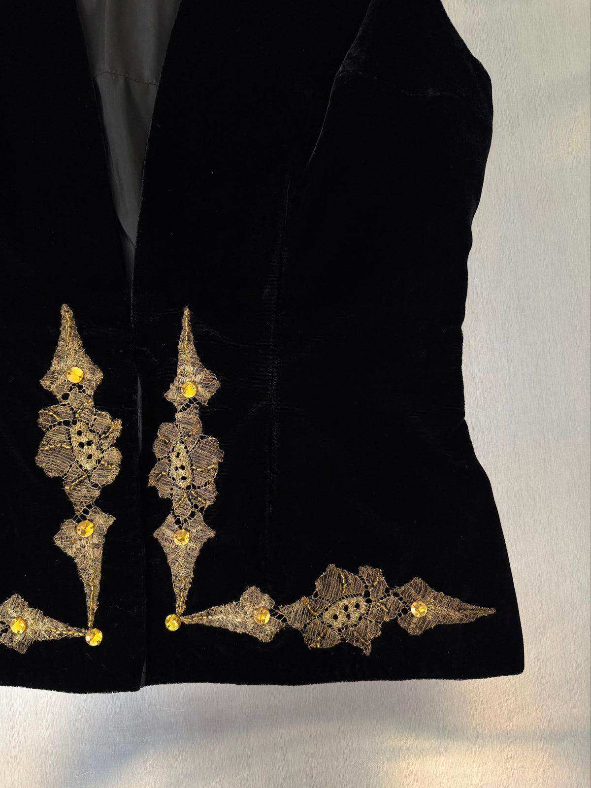 Close-up of a black velvet vest with ornate gold lace embroidery and small yellow bead accents along the front opening.