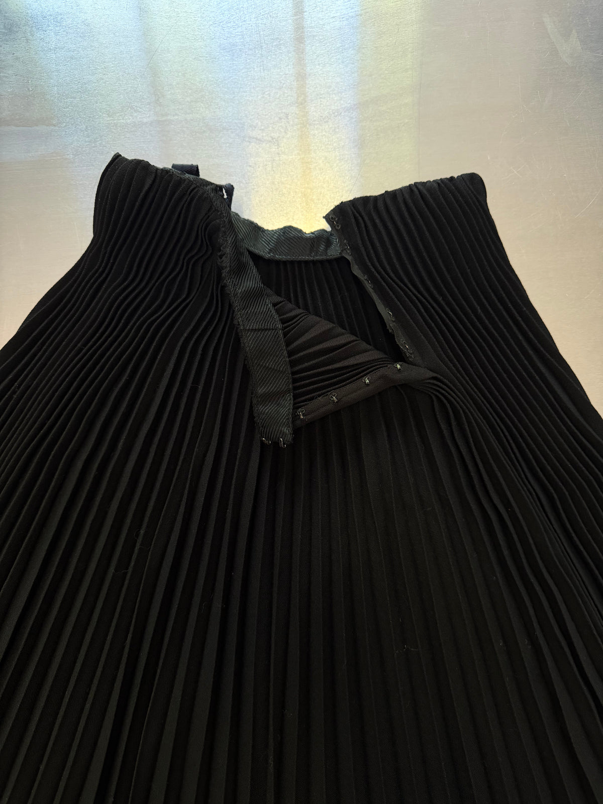 A close-up of a black pleated garment, likely a pleated dress or long skirt, featuring a structured shoulder and a V-shaped neckline with a black fabric trim detail.