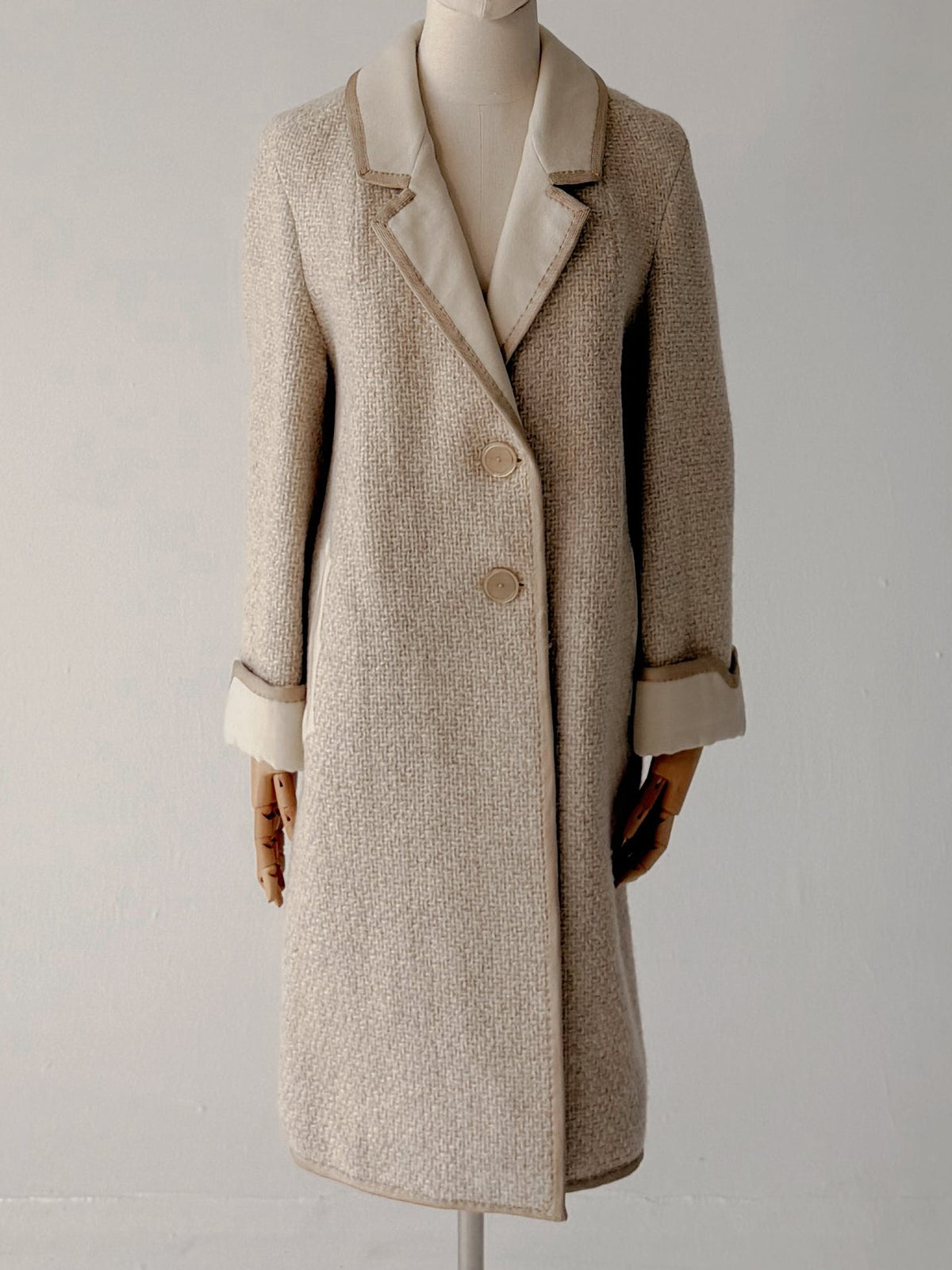 A light beige wool-blend coat displayed on a headless mannequin stand against a plain off-white studio background.