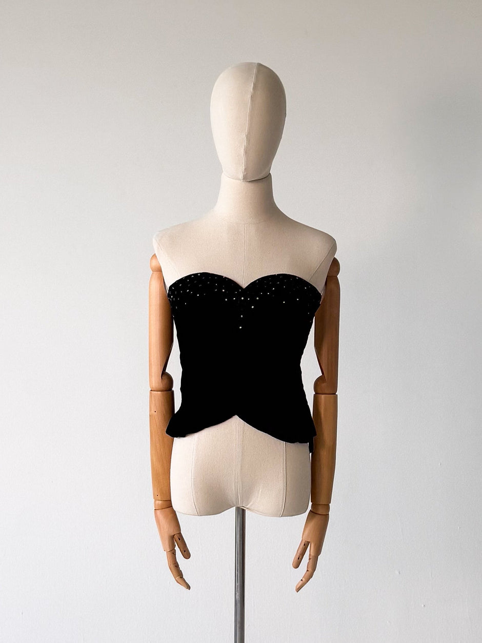 A black strapless corset-style top displayed on a beige mannequin torso with wooden arms, mounted on a silver stand against a plain light gray studio wall.