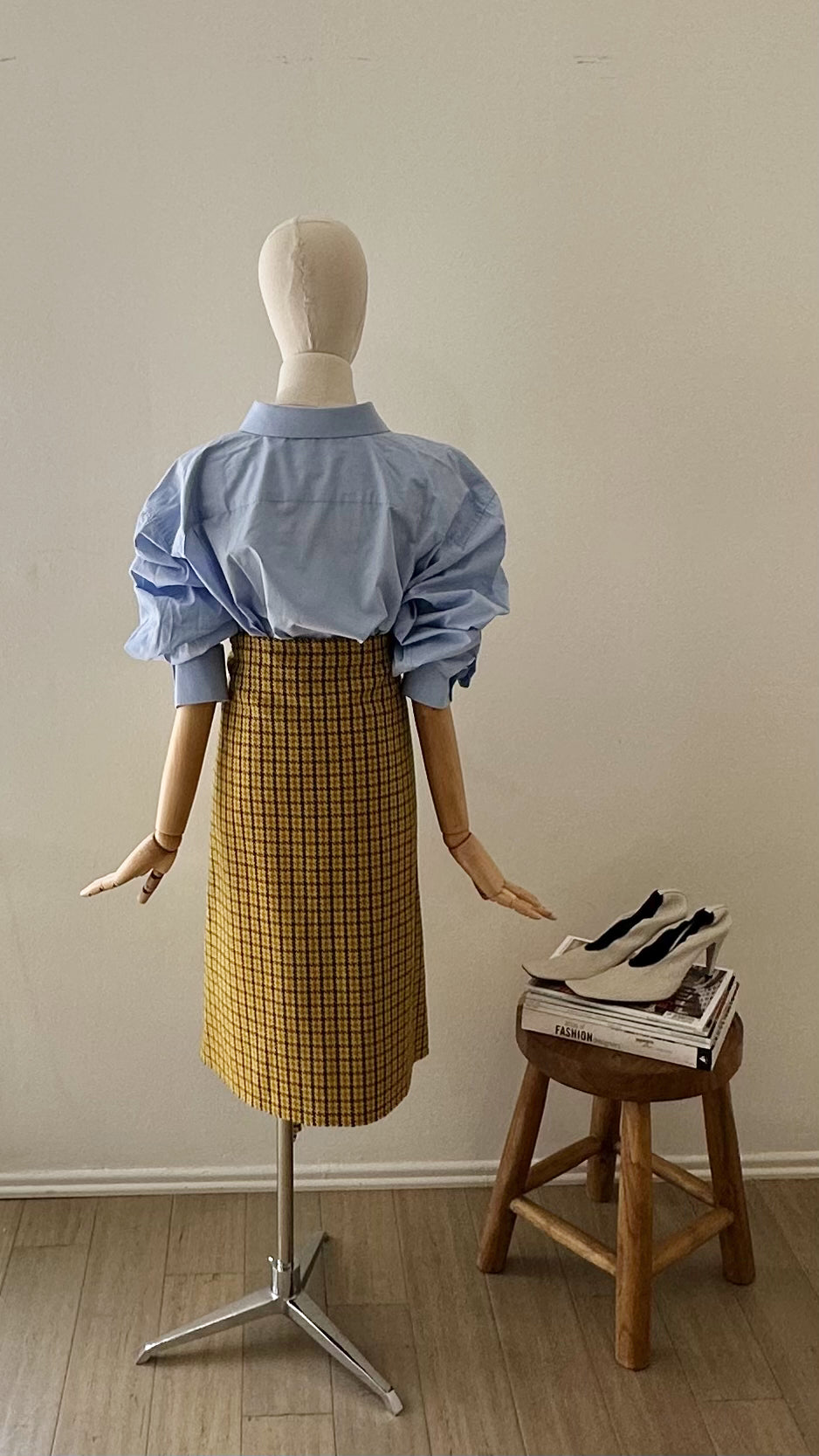 Mannequin wearing a blue shirt and Balenciaga yellow plaid skirt next to a wooden stool with shoes on a neutral background