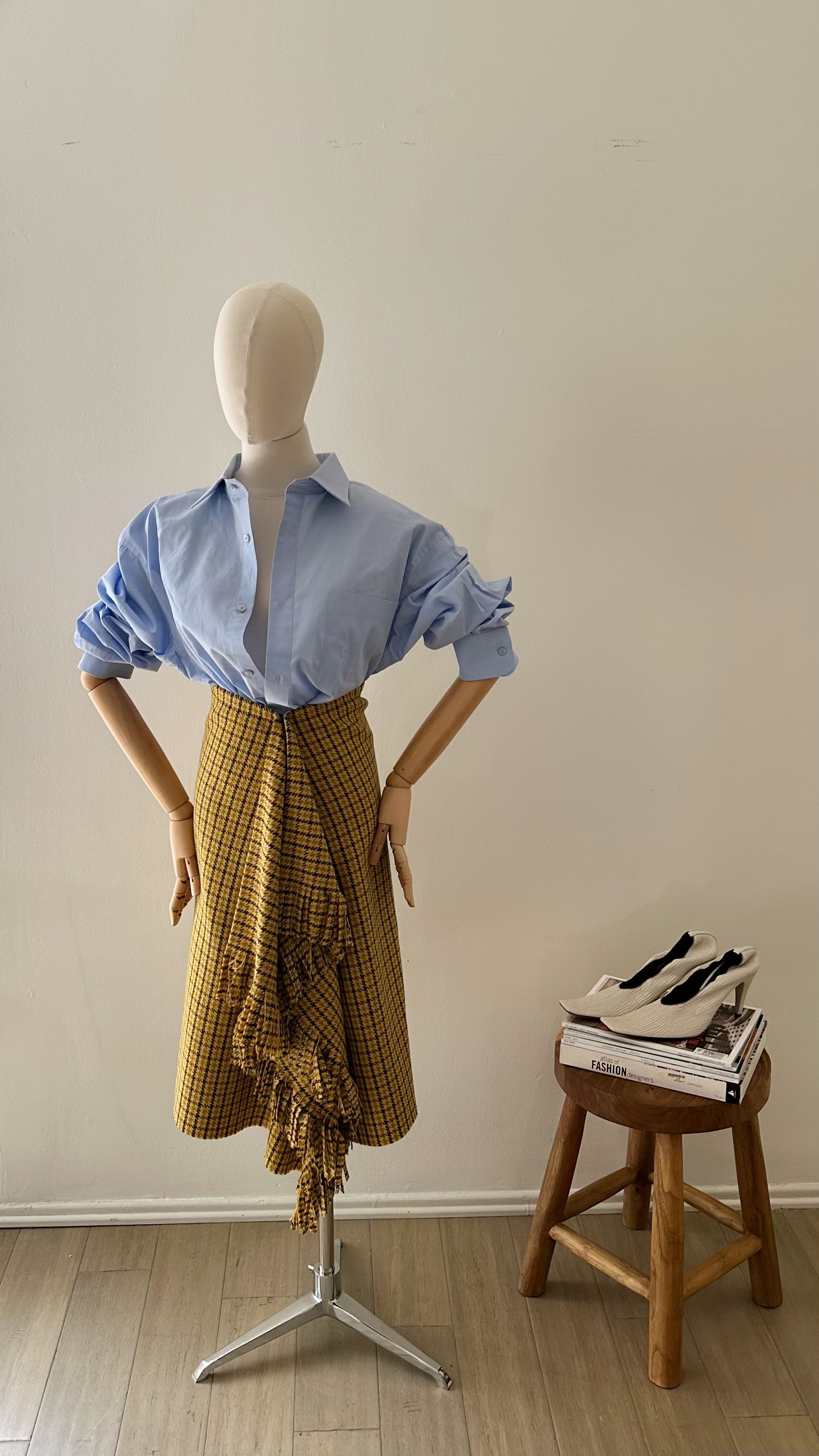 Mannequin wearing a blue shirt and yellow plaid skirt with a wooden stool and shoes in the background.