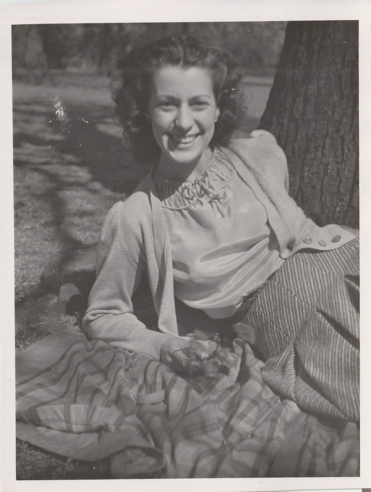 Black and white vintage portrait of a smiling woman sitting outdoors against a tree trunk, wearing a light blouse and cardigan with a striped skirt.