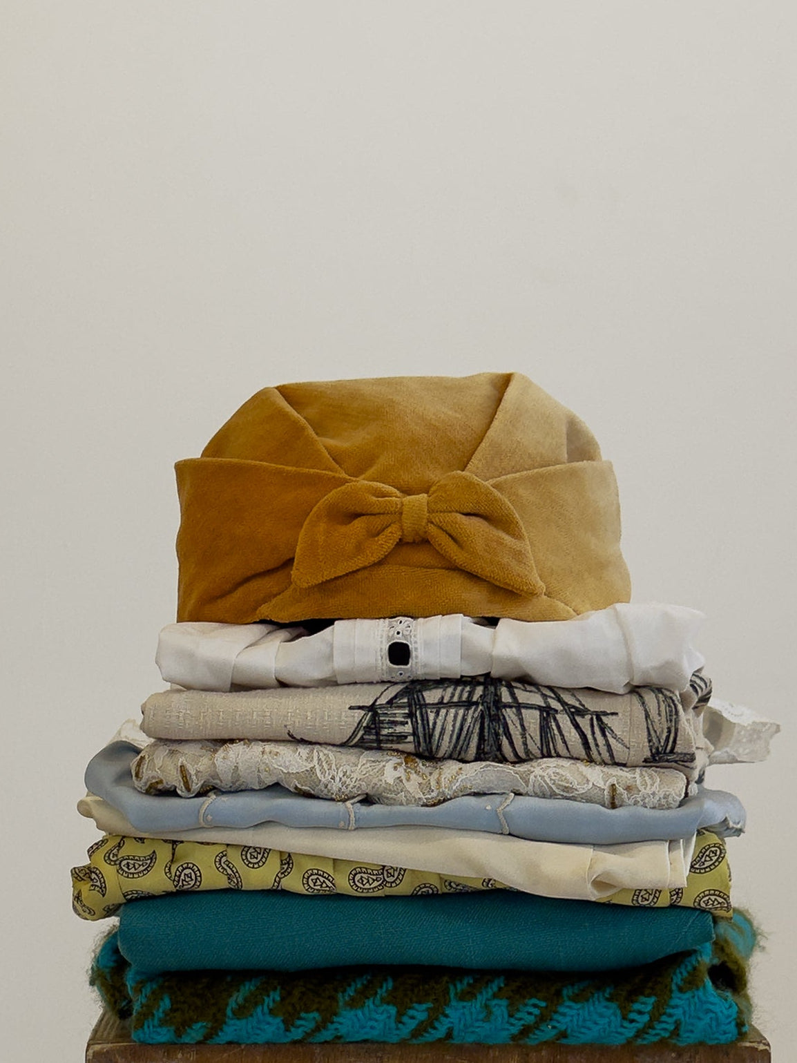 A stack of folded patterned and solid-colored garments sits on top of a wooden block against a plain light beige studio background.