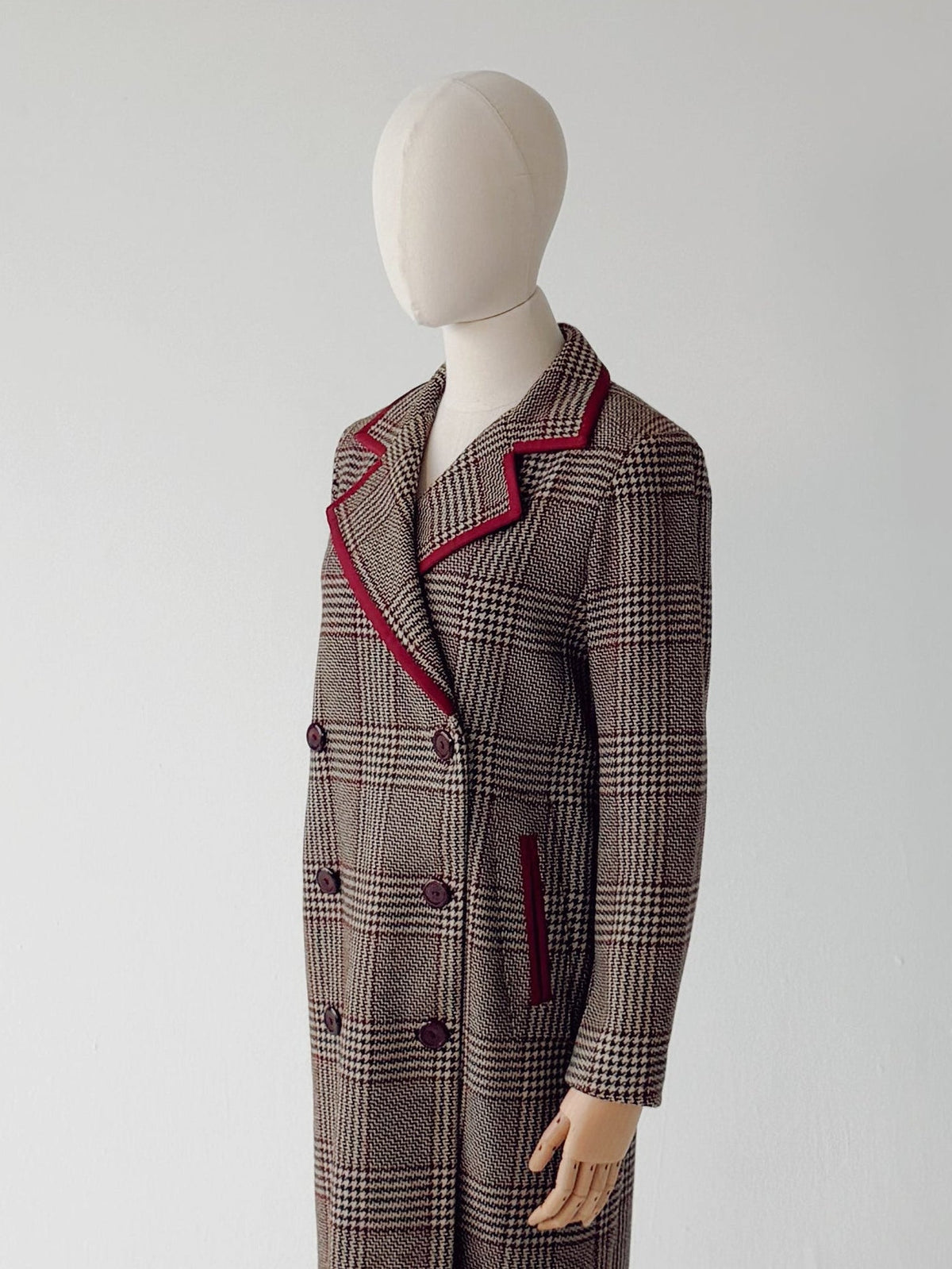 A long double-breasted plaid coat displayed on a mannequin stand against a plain light gray studio background. The coat has a brown and gray plaid pattern with red piping along the lapels and pocket edges.