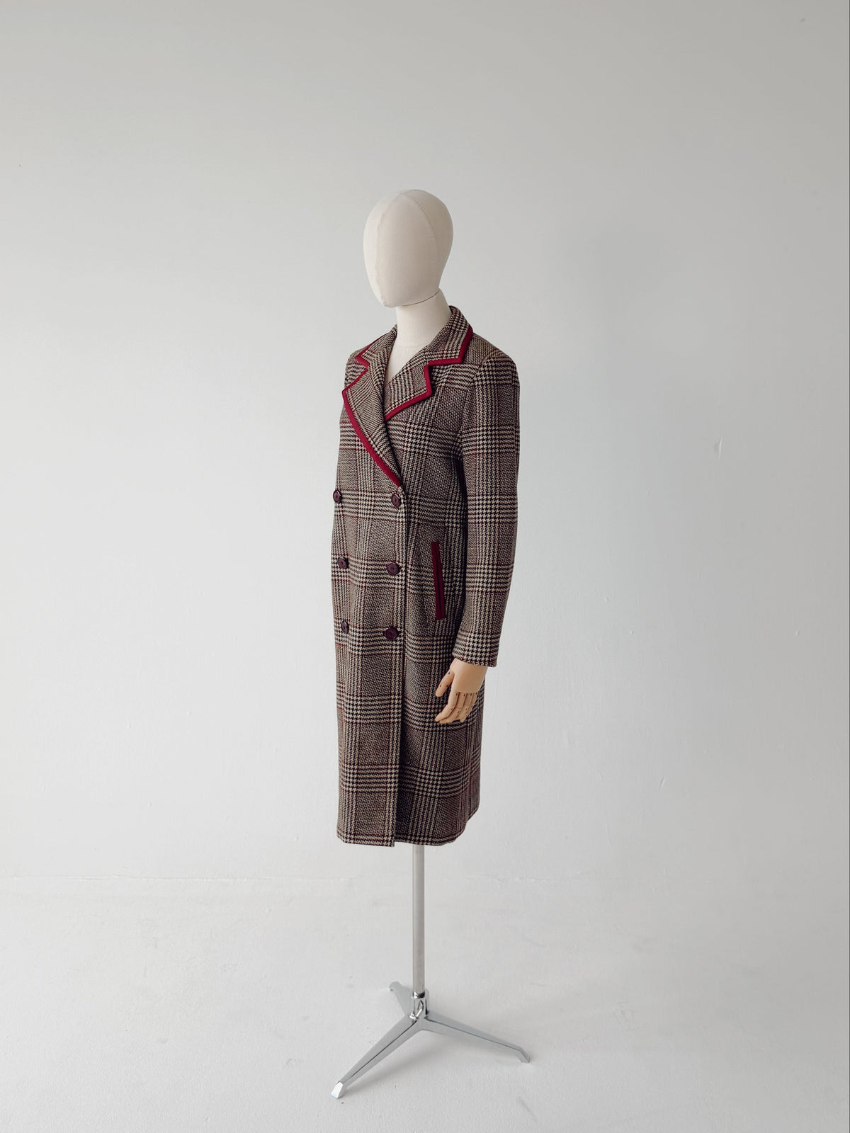 A long double-breasted plaid coat displayed on a mannequin stand against a plain light gray studio background. The coat has a brown and gray plaid pattern with red piping along the lapels and pocket edges.