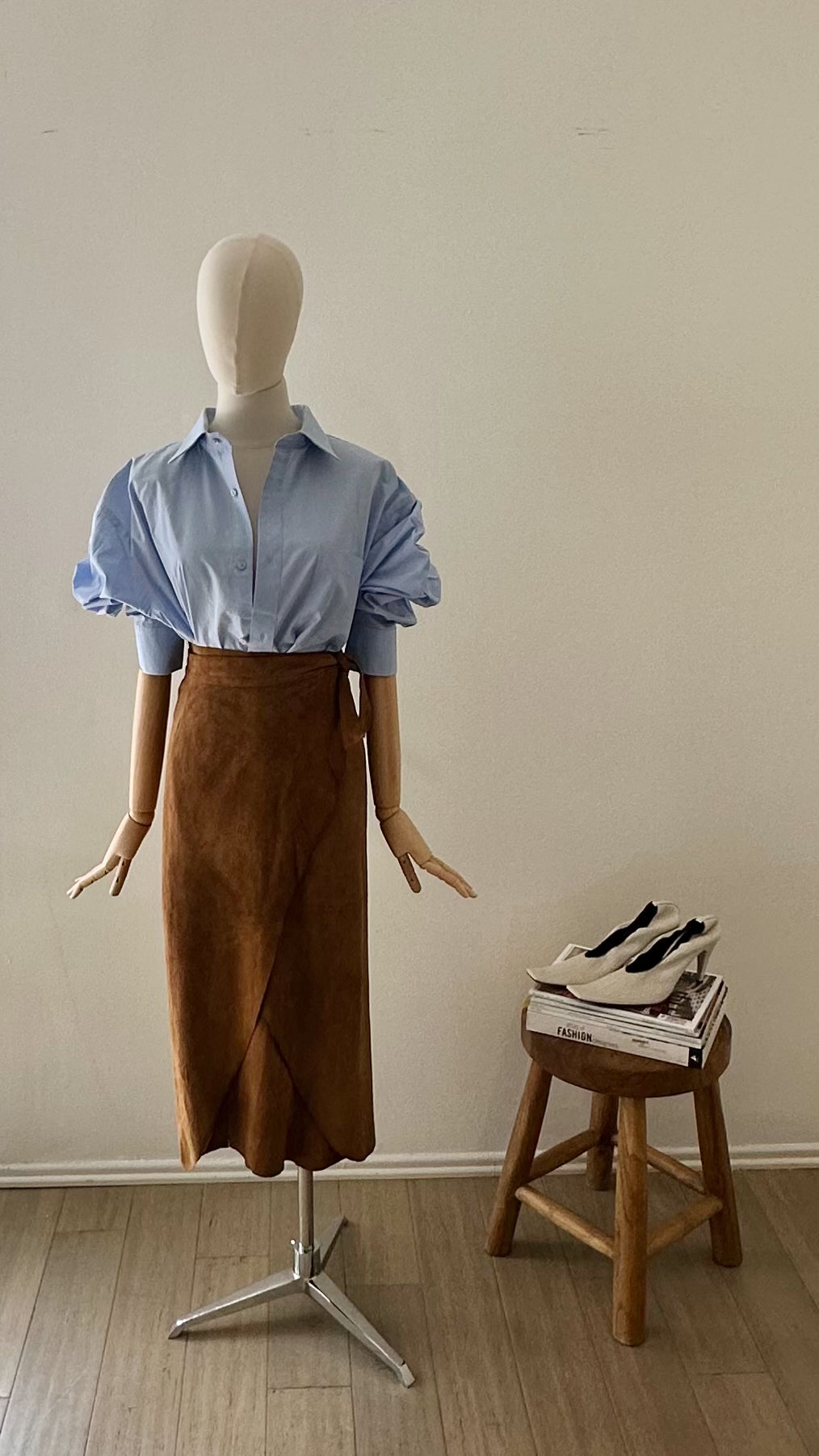 Mannequin wearing a blue shirt and brown skirt next to a wooden stool with books on it against a plain wall.